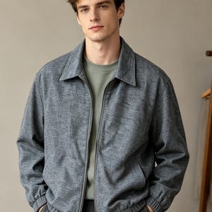May include: A gray zip-up jacket featuring a subtle textured pattern, a pointed collar, and long sleeves with elasticized cuffs. The jacket is worn over a solid light green crew neck shirt and paired with dark gray trousers.