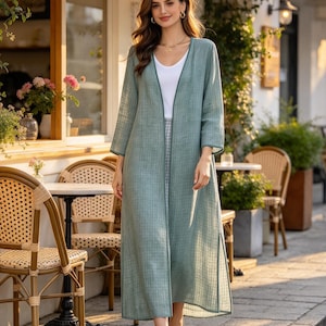 May include: A long, sage green open-front duster cardigan featuring a subtle grid pattern, three-quarter length sleeves, and side slits. The garment is worn over a simple white V-neck top and light-colored trousers, creating a layered look.
