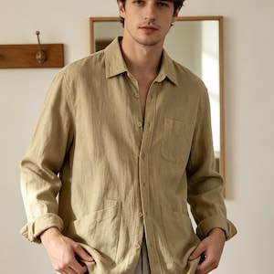 May include: A long-sleeved button-down shirt in a light olive green colour. The shirt features a classic collar, a single chest pocket on the left side, and two larger patch pockets at the bottom front. The fabric has a visible textured weave.