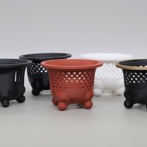 May include: A collection of five small, round, footed pots with openwork lattice patterns on the sides. The pots are available in matte black, terracotta red, and white, each featuring three rounded feet and a wide, circular rim for planting.