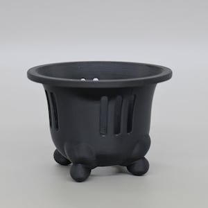 May include: A matte black plant pot featuring a wide, circular rim and a series of vertical rectangular cutouts along the sides. The container is supported by three rounded, bulbous feet at the base, providing a stable foundation for the vessel.