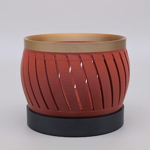 May include: A cylindrical planter featuring a red body with diagonal cutouts, a metallic gold rim at the top, and a solid black circular base. The structure has a layered texture and a wide opening, designed to hold small plants or decorative items.