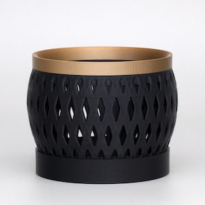 May include: A cylindrical planter featuring a matte black body with a repeating diamond-shaped cutout pattern. The top rim is finished in a metallic gold tone, and the base consists of a solid black circular stand that supports the main structure.