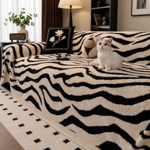 Puede incluir: Una funda de sofá de felpa con un patrón de rayas de cebra en negro y crema. La tela texturizada cubre el mueble, acompañada por un cojín cuadrado negro con bordado floral blanco y un pequeño cojín redondo en tonos beige y marrón.