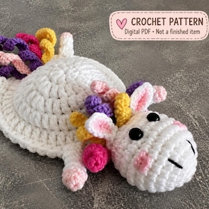 Puede incluir: Un unicornio de ganchillo blanco con cuerpo plano y circular, cabeza con ojos negros y una melena y cola multicolor hechas de espirales de hilo amarillo, rosa y morado. Un cuadro de texto dice: CROCHET PATTERN Digital PDF Not a finished item.