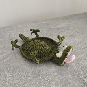 Puede incluir: Un posavasos de ganchillo color verde oliva con forma de caimán tumbado. El caimán tiene un cuerpo redondo, cuatro patas pequeñas, una cola y una cabeza con ojos blancos, pupilas negras y la boca abierta mostrando una lengua y el interior rosa.