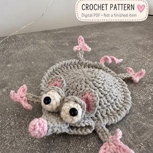 Puede incluir: Una criatura de ganchillo gris con cuerpo redondo, cuatro extremidades con puntas rosas y nariz rosa. Tiene dos ojos grandes blancos con pupilas negras y orejas pequeñas con borde rosa. El texto dice: CROCHET PATTERN Digital PDF.