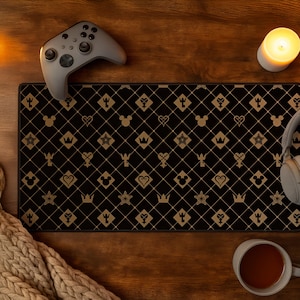Puede incluir: Una alfombrilla de escritorio rectangular con fondo negro y un patrón repetitivo de diamantes dorados. El diseño incluye iconos dorados de cabezas de Mickey Mouse, coronas, estrellas, corazones y símbolos en forma de llave en los diamantes.