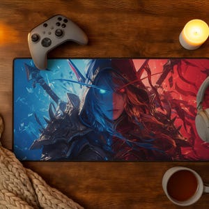 Può includere: Un tappetino per mouse rettangolare da gioco con un'illustrazione bicolore di un personaggio incappucciato con un occhio blu brillante e uno rosso brillante, che indossa un'armatura scura. Il tappetino è circondato da un controller, cuffie e una candela.