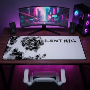 May include: A rectangular desk mat featuring a white background with a black ink-style splatter design on the left side, a circular occult symbol, and the text "SILENT HILL" printed in black on the right side, with a black stitched border.