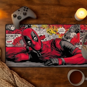 May include: A rectangular desk mat featuring a comic book style illustration of the character Deadpool in a red and black suit, lying down against a background of action-oriented comic panels with various sound effects and speech bubbles.