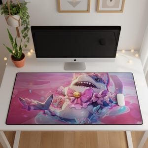 Puede incluir: Una alfombrilla de escritorio rectangular con una colorida ilustración de un tiburón de dibujos animados que lleva un traje rosa con volantes, un lazo y una pequeña campana. El tiburón tiene una amplia sonrisa sobre un fondo rosa suave.
