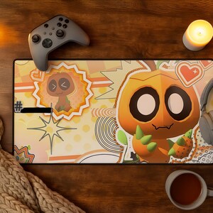 May include: A rectangular desk mat featuring a large orange cartoon character with big eyes and green claws, surrounded by smaller graphic elements like hearts, stars, and a smaller character in a sunburst frame, all set against a light yellow background.