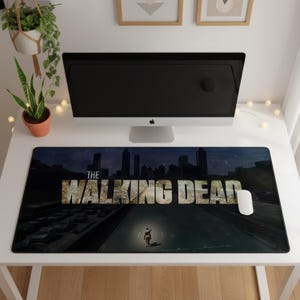 May include: A rectangular desk mat featuring a dark, atmospheric scene from The Walking Dead. The graphic displays a city skyline at night, a highway filled with abandoned cars, and a lone figure on horseback, with the title text prominently displayed.