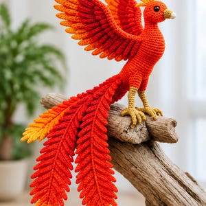 Könnte beinhalten: Eine gehäkelte Phönix-Figur aus leuchtend rotem, orangefarbenem und gelbem Garn. Der Vogel hat ausgebreitete Flügel, einen langen, gefiederten Schwanz und strukturierte goldgelbe Beine und sitzt auf einem Stück Treibholz.