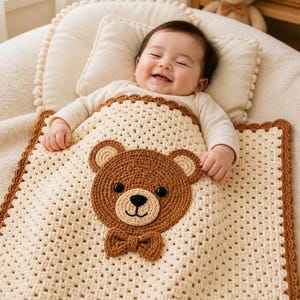 Könnte beinhalten: Eine quadratische Häkeldecke für Babys in Creme mit braunem Rand, die in der Mitte eine große, aufgesetzte braune Bärengesicht-Applikation mit schwarzen Knopfaugen, einer gestickten Nase und einer kleinen Fliege als Detail aufweist.