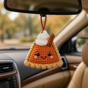 Puede incluir: Un adorno colgante de ganchillo con forma de porción de tarta de calabaza, con corteza color canela, relleno naranja, nata montada blanca y una rama de canela marrón. La tarta tiene una cara sonriente con ojos negros y mejillas rosadas.