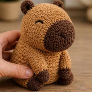 Pode incluir: Um pequeno brinquedo de pelúcia de capivara feito de crochê, de formato redondo, com corpo castanho claro, focinho, orelhas e patas castanho escuro. O brinquedo tem olhos pretos bordados e um nariz simples costurado, com acabamento em fio.