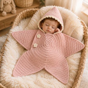 Peut inclure: Un bébé portant une couverture d'emmaillotage en crochet rose poudré en forme d'étoile avec une capuche pointue. La couverture présente une bordure festonnée crème le long de la capuche et deux boutons en forme de fleur de couleur crème.