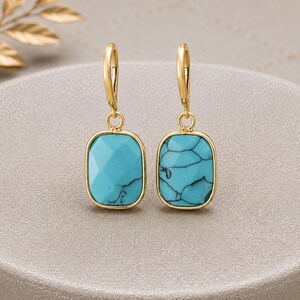 Puede incluir: Un par de pendientes colgantes de tono dorado con piedras rectangulares de color turquesa con vetas oscuras. Cada piedra está engastada en un marco de metal liso de color dorado y cuelga de un cierre de gancho sencillo de tono dorado.