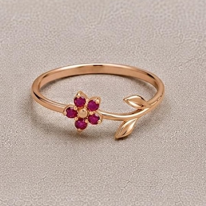Può includere: Un anello in oro rosa caratterizzato da un delicato motivo floreale composto da cinque piccole pietre preziose rotonde di colore rosa vivace disposte a fiore, con una fascia sottile che termina con un dettaglio a foglia in metallo lucido.