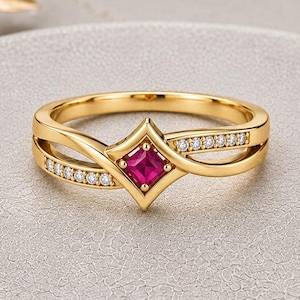 May include: A gold ring featuring a central princess-cut vibrant pink gemstone set within a diamond-shaped frame. The band splits into two delicate arms, each accented with a row of small, clear round-cut stones that extend along the sides.