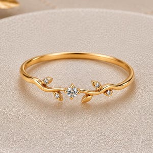 May include: A gold ring featuring a delicate vine-like band with small leaves and four embedded clear stones. The central stone is slightly larger than the three smaller stones positioned along the curved, branch-inspired metal band.