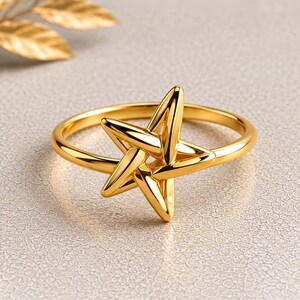May include: A polished gold ring featuring a prominent five-pointed star design centered on a thin, smooth circular band. The star has rounded, tapered points and a reflective, metallic finish that catches the light, creating bright highlights.