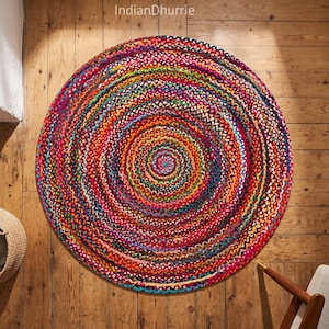 May include: A round, braided rug featuring a vibrant, multi-colored pattern with concentric circles of red, orange, yellow, green, blue, and purple fibers. The text IndianDhurrie is visible at the top center of the image above the rug.