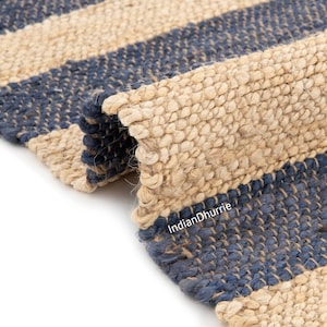 Puede incluir: Una alfombra tejida con rayas horizontales alternas en beige natural y azul marino intenso. La textura es gruesa y fibrosa, mostrando un patrón de tejido trenzado. El texto IndianDhurrie está impreso en blanco sobre la superficie.