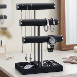 May include: A black velvet jewelry display stand with three horizontal bars at varying heights for necklaces, bracelets, and a watch. The base features a rectangular tray with slots for rings and a flat section for small items like keys or accessories.