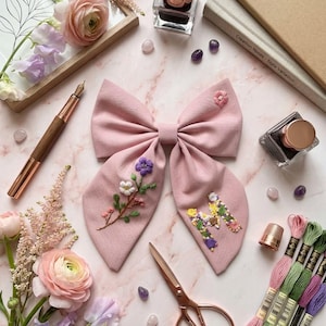 Può includere: Un fiocco per capelli in tessuto rosa polvere con dettagli floreali ricamati a mano sul lembo sinistro e una lettera M a motivi floreali sul lembo destro. Il fiocco è realizzato in tessuto testurizzato e presenta un piccolo fiore ricamato.
