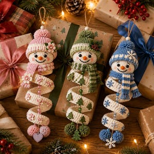 May include: Three crocheted snowman ornaments with spiral bodies. The left snowman wears a pink hat and scarf with flower accents. The center snowman has a green hat and scarf with tree motifs. The right snowman features a blue hat and scarf with snowflake details.