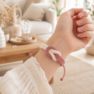 May include: A macrame bracelet featuring a central knot design, crafted from intertwined cream and dusty rose colored cord. The bracelet includes adjustable pull strings with small knotted ends, creating a simple and functional wrist accessory.
