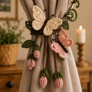 May include: A crochet curtain tieback featuring a large cream butterfly and a smaller pink butterfly, accented with green crocheted leaves and pink flower buds on brown stems, secured around a solid light beige fabric curtain panel.