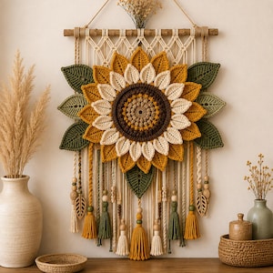 May include: A wall hanging featuring a large crocheted sunflower with yellow, cream, and brown petals. The piece is suspended from a wooden dowel with macrame cord, green crocheted leaves, wooden beads, and dangling tassels in shades of green, yellow, and cream.