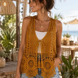 Può includere: Un gilet senza maniche all'uncinetto color giallo senape con un intricato motivo traforato, un orlo smerlato e una chiusura frontale a laccio con due nappe abbinate. Il gilet è indossato sopra una canotta bianca e pantaloncini di jeans azzurri.