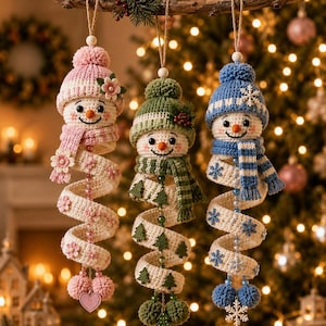 May include: Three hanging crochet snowman ornaments with spiral bodies. The left snowman wears a pink hat and scarf with flower accents. The center snowman has a green hat and scarf with tree motifs. The right snowman features a blue hat and scarf with snowflake details.