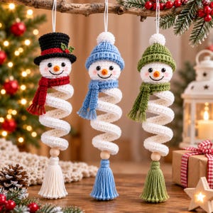 May include: Three crocheted snowman ornaments hanging vertically. Each features a white spiral body, a wooden bead, and a matching tassel. The first wears a black top hat and red scarf, the second a blue beanie and scarf, and the third a green beanie and scarf.
