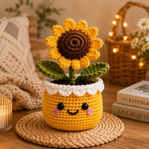 Pode incluir: Um girassol de crochê num vaso amarelo com um rosto sorridente, olhos pretos e bochechas cor-de-rosa. O girassol tem um centro castanho escuro, pétalas amarelas brilhantes e duas folhas verdes. O vaso tem um rebordo branco recortado.