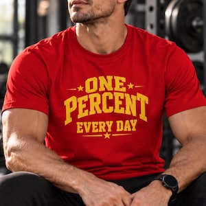 Könnte beinhalten: Ein rotes kurzärmeliges T-Shirt mit Rundhalsausschnitt und dem gelben Schriftzug "ONE PERCENT EVERY DAY" auf der Brust in einer fetten, sportlichen Schriftart, akzentuiert durch kleine Sterne.