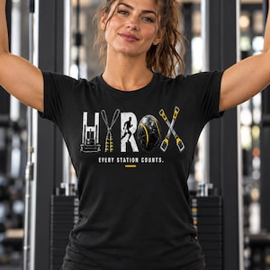 Könnte beinhalten: Ein schwarzes Kurzarm-T-Shirt mit dem weißen Schriftzug HYROX, in dessen Buchstaben Symbole für Fitnessgeräte integriert sind. Unter dem Text steht der Satz EVERY STATION COUNTS in Weiß mit einer kleinen gelben Linie darunter.