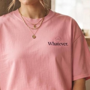 Pode incluir: Uma t-shirt de manga curta cor-de-rosa clara com o texto "Whatever." impresso em letras escuras na parte superior esquerda do peito, acompanhado por um pequeno gráfico oval com as letras "UM" acima da palavra.