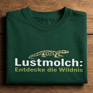 Op de afbeelding: Een donkergroen t-shirt met een afbeelding van een lichtgekleurde hagedis gecentreerd boven de witte tekst "Lustmolch:" en de lichtgroene tekst "Entdecke die Wildnis" gedrukt op de voorzijde in een strak, schreefloos lettertype.