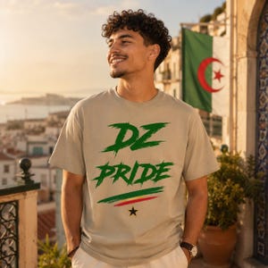 Könnte beinhalten: Ein beiges kurzärmeliges T-Shirt mit dem grünen Schriftzug DZ PRIDE in Pinselstrich-Optik, ergänzt durch einen kleinen horizontalen grün-gelb-roten Streifen über einem schwarzen Stern, der mittig unter dem Text auf der Vorderseite platziert ist.