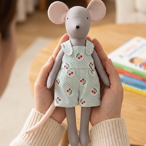 May include: A plush mouse toy with grey fabric skin, large rounded ears, and a long tail. The mouse is wearing light green overalls featuring a white polka dot pattern and a repeating red cherry print with green stems and leaves.
