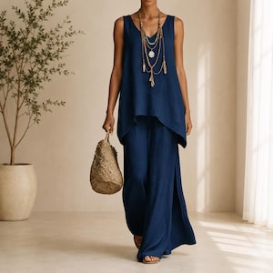 May include: A dark blue sleeveless tunic top and matching wide-leg trousers with side slits. The outfit is paired with a layered gold-tone necklace featuring a turquoise bead and a textured, woven straw handbag with a rounded shape.