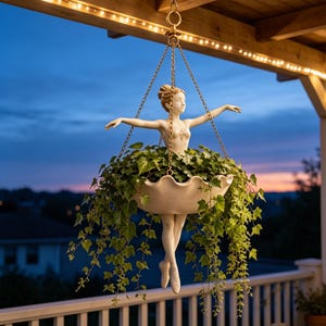 Peut inclure: Une jardinière suspendue représentant une ballerine aux bras étendus. La figurine porte un corsage blanc et des chaussons de danse, avec une jupe en forme de bol festonné contenant du lierre vert. Elle est suspendue par des chaînes dorées.