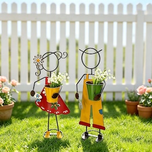 May include: Two metal garden stakes shaped like stylised figures. The left figure wears a red dress with green floral accents and holds a yellow bucket. The right figure wears yellow overalls and holds a green bucket. Both contain white flowers.