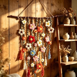 Pode incluir: Uma tapeçaria de parede com vários fios verticais de trepadeiras de crochê adornadas com várias flores de lã coloridas em tons de vermelho, laranja, rosa, azul, branco e amarelo, suspensa por um ramo de madeira natural com uma corda.